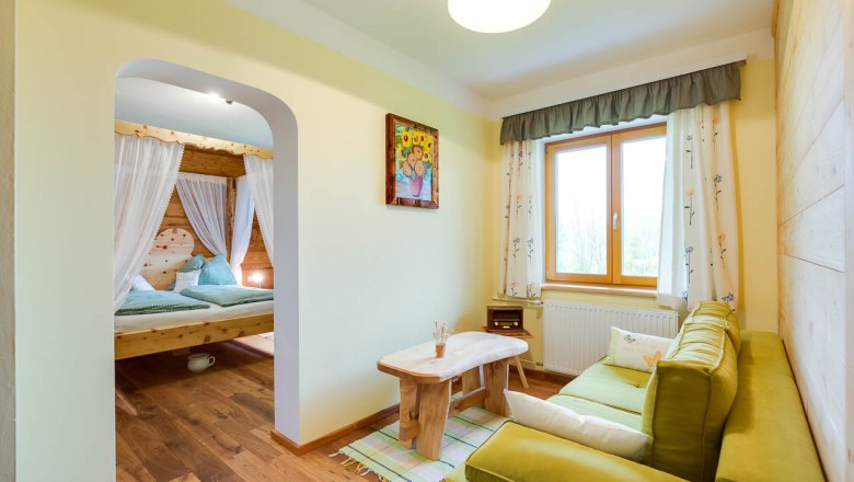 Zimmer im Höchbauernhof, © zVg Gemütliches Zimmer mit Holzmöbeln, grünem Sofa, großem Fenster und einem Himmelbett im Hintergrund.