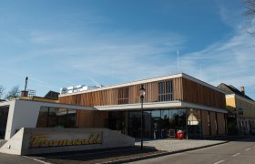 Der Hubertushof in Bad Fischau-Brunn, © Fotografin: Eva Heindl Modernes Gebäude mit Holzfassade und Glasfront, davor ein Schild mit der Aufschrift 'Fromwald'.