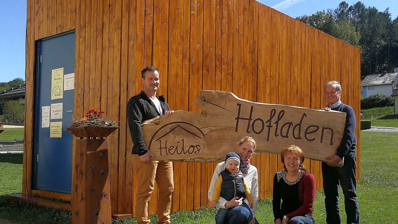 Ab Hof Verkauf Heilos-Aussenansicht, © Claudia Gugler Familie vor einem Holzgebäude mit einem Schild 'Heilos Hofladen'.