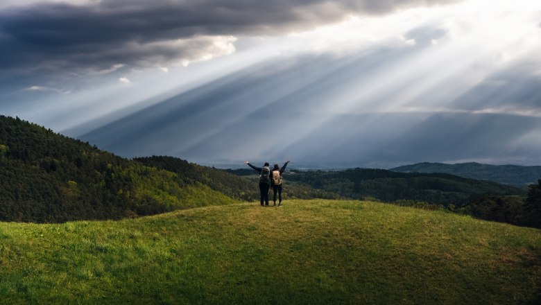 Lebensweg, © Waldviertel Tourismus, Melanie Többe Zwei Personen stehen auf einem Hügel mit Rucksäcken, umgeben von grünen Wäldern und dramatischen Sonnenstrahlen durch Wolken.