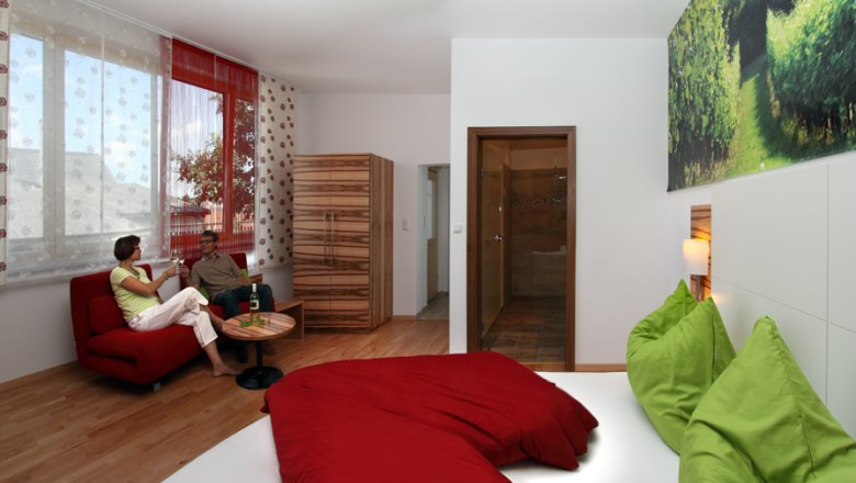 Gästezimmer, © Familie Bohrn Modernes Gästezimmer mit rotem Sofa, grün-rotem Bett und großem Fenster.