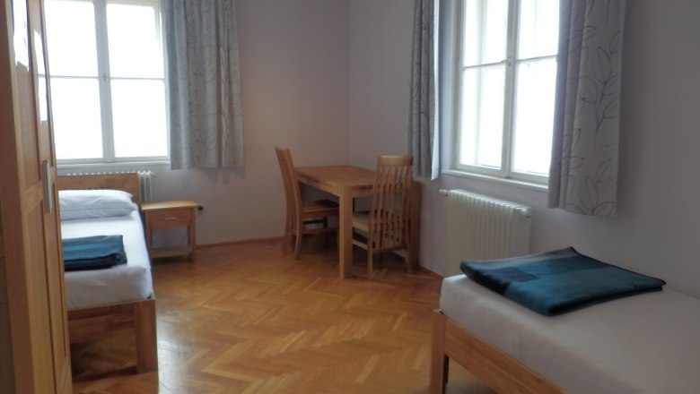 Gästezimmer, © Geistliches Jugendzentrum Oberleis Ein einfach eingerichtetes Gästezimmer mit zwei Betten, einem Tisch und Stühlen, Holzboden und großen Fenstern mit Vorhängen.
