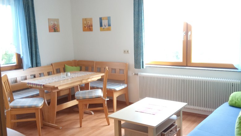 FW "Panoramablick", © Blumentalhof Helles Wohnzimmer mit Holzmöbeln, Eckbank, Tisch, Sofa und großen Fenstern mit blauen Vorhängen.