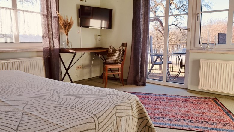 Zimmer im Haupthaus, © Szilágyi Gemütliches Zimmer mit Bett, Tisch, Stuhl und Fernseher. Große Fenster mit Blick auf Terrasse und Bäume. Teppich auf dem Boden.