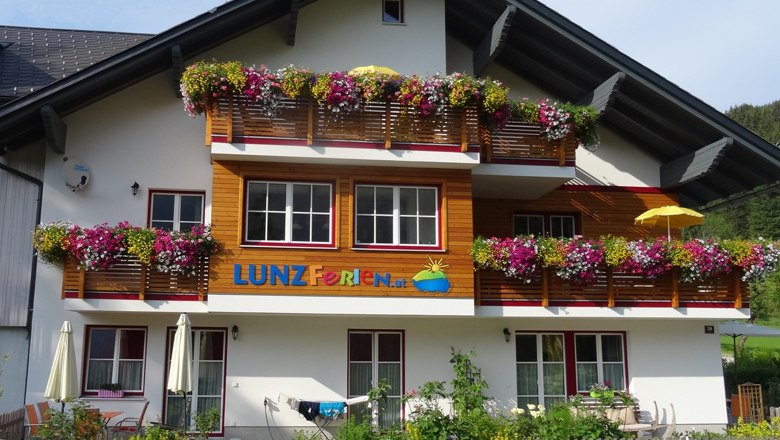 Lunzferien Geli Mayr, © zVg Ein Ferienhaus mit bunten Blumen auf den Balkonen und dem Schriftzug 'LUNZ Ferien'.