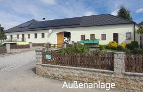 Außenanlage – Ferienwohnung rechts, © Erika Polt Ein großes Gebäude mit gelber Fassade und schwarzem Dach, umgeben von einem Garten und einem Holzzaun. Der Himmel ist blau mit einigen Wolken.