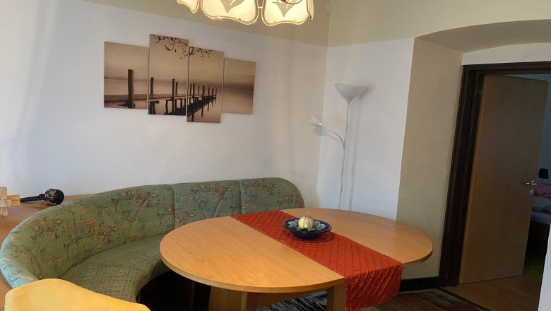 Wohnzimmer, © Wiener Alpen Ein Wohnzimmer mit einem ovalen Holztisch, einer grünen Eckbank, einem roten Tischläufer und einem Bild an der Wand. Eine Stehlampe steht in der Ecke.