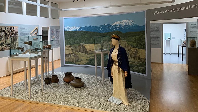 PIZ1000 - Pittener Regionsmuseum, © Marktgemeinde Pitten Ausstellung im Pittener Regionsmuseum mit historischen Artefakten und einer Figur in traditioneller Kleidung vor einem Wandbild.