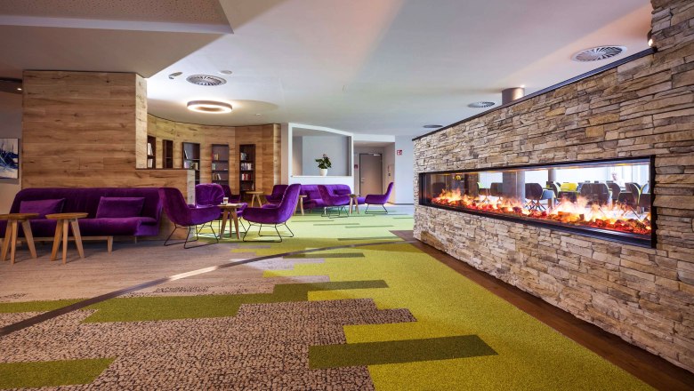 Lobby, © Hannes Dabernig Moderne Hotellobby mit grün-braunem Teppichboden, Kamin, lila Sofas und Stühlen und Holztischen.