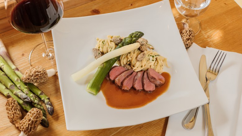 Kaiser von Österreich, © Rita Newman Gourmetgericht mit Fleisch, Spargel, Nudeln und Morcheln auf einem weißen Teller, daneben ein Glas Rotwein.