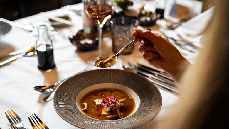 Das Linsberg - Restaurant, © Linsberg Asia Eine Person hält einen Löffel über einem Teller mit Suppe auf einem gedeckten Tisch.
