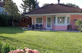 Haus Muck, © Barbara Muck Ein rosa Haus mit Garten, Terrasse und Gartenmöbeln, umgeben von Bäumen und Blumen.