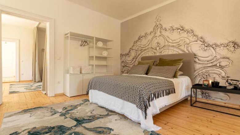 Beispiel Schlafbereich, © Guestia GmbH Ein stilvoll eingerichtetes Schlafzimmer mit einem großen Bett, dekorativen Kissen, einem Teppich und einer kunstvollen Wandgestaltung.