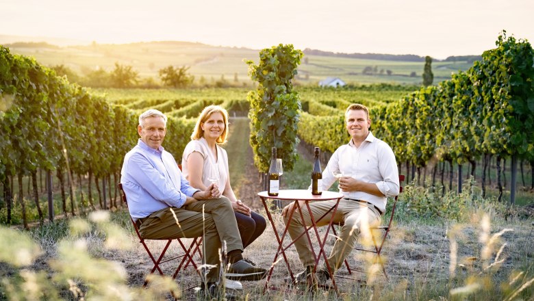 Winzerfamilie Stift, © Substudio Paukner Drei Personen sitzen an einem Tisch im Weinberg mit Weingläsern und Flaschen.