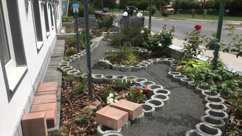 Rosengarten, © Familie Mitra Ein kleiner Garten mit Rosen und einem geschwungenen Weg aus Betonsteinen neben einem Gebäude.