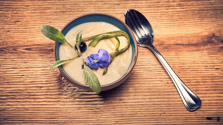 Spargelcremesuppe, © Niederösterreich Werbung/Andreas Hofer Spargelcremesuppe Suppe mit Blüte und Kräutern auf Holztisch, Löffel daneben.