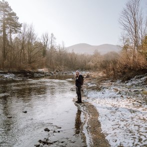 Wandern durch das winterliche Helenental, © Niederösterreich Werbung/Robin Uthe Junge Frau steht auf Flussbett, dahinter kahle Winterlandschaft in sanftem Winterlicht.