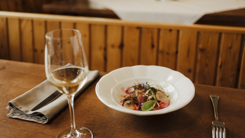 Regionale Weinauswahl, © Niederösterreich Werbung/Michael Reidinger Ein Glas Weißwein neben einem Teller mit einem dekorativen Gericht auf einem Holztisch.