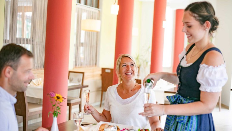 Mittagessen im Wintergarten, © Schwarz-König/Sinzinger – www.sksfoto.at Eine Frau in Tracht schenkt einem Paar in einem Restaurant Sekt ein.