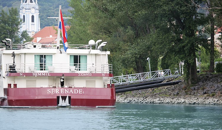 Anlegestelle Dürnstein 22 mit Boot, © Donau Schiffsstationen GmbH Ein Flusskreuzfahrtschiff namens Serenade an einer Anlegestelle in Dürnstein, mit einem Kirchturm im Hintergrund.