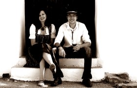 Winzer, © Weingut A+A Mayer Ein Mann und eine Frau sitzen auf einer Treppe, der Mann hält ein Weinglas.