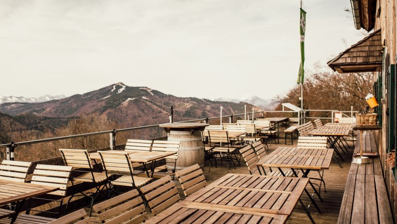 Von der Terrasse, © Stefanie Rysavi Leere Terrasse mit Holztischen und -stühlen, Berglandschaft im Hintergrund.