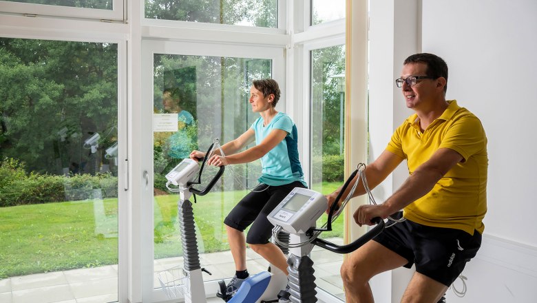 Fahrradergometertraining, © HKZGG, Robert Herbst Zwei Personen trainieren auf Fahrradergometern in einem Raum mit großen Fenstern.