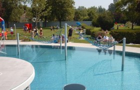 Erlebnisbad "Aqua Splash", © Erlebnisbad "Aqua Splash" Ein Freibad mit Hängematten über dem Wasser und Menschen im Hintergrund auf der Liegewiese.