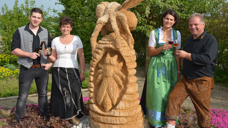 Hannes, Bettina, Astrid und Alfred, © Imkerei Stögerer, Fotograf Gerhard Kapeller Vier Personen in traditioneller Kleidung stehen neben einer Holzskulptur im Freien.