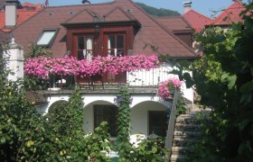 Gästehaus Haiminger, © Familie Schmelz Ein Gästehaus mit einem Balkon voller rosa Blumen, umgeben von grüner Vegetation.