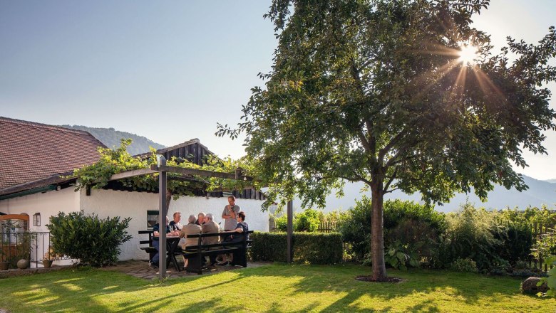 beisammensitzen-im-garten_c_urbanushof-_-leonhard-hilzensauer_preview, © Leonhard Hilzensauer Menschen sitzen an einem Tisch im Garten unter einer Pergola, umgeben von Natur und Sonnenschein.