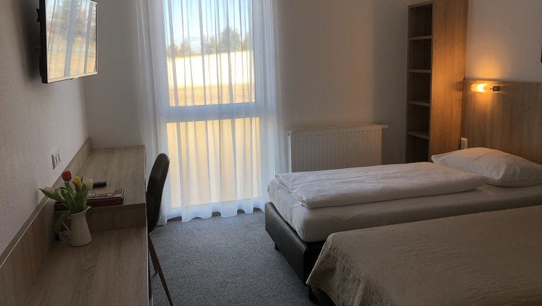 Bernstein Motel, © Bernstein Motel Ein schlicht eingerichtetes Motelzimmer mit zwei Einzelbetten, einem Schreibtisch, einem Fernseher und einem Fenster mit Vorhängen.