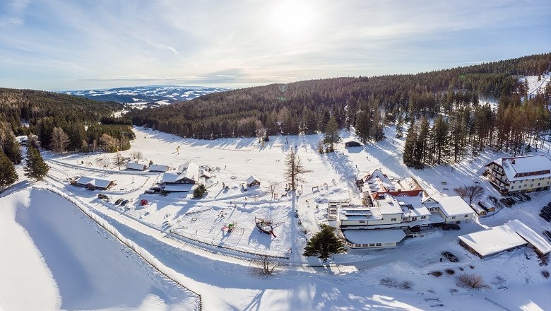 Schigebiet Mönichkirchen-Mariensee, © Skiregion Ostalpen/Martin Fülöp Luftaufnahme eines verschneiten Skigebiets mit Gebäuden und Wald im Hintergrund.