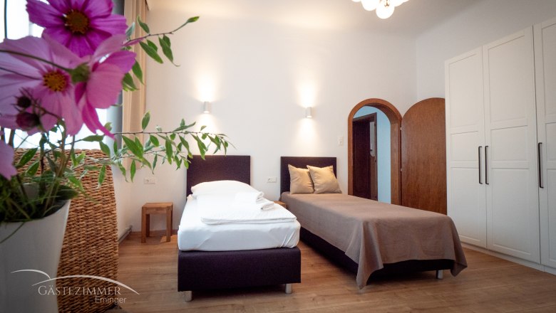 Zweibettzimmer, © Doris Eminger Ein gemütliches Zweibettzimmer mit Blumen im Vordergrund, zwei Betten, einem Schrank und einer Lampe an der Decke.