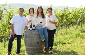 TDR-Heuriger Familie Kienberger, © Kienberger Vier Personen stehen und sitzen in einem Weinberg, lächelnd mit Weingläsern in der Hand, neben einem Weinfass mit Flaschen darauf.