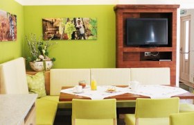 Gästehaus Loiskandl, © Gästehaus Loiskandl / Elisabeth Widhalm Gemütlicher Essbereich mit grünen Wänden, Holzmöbeln und einem Fernseher.