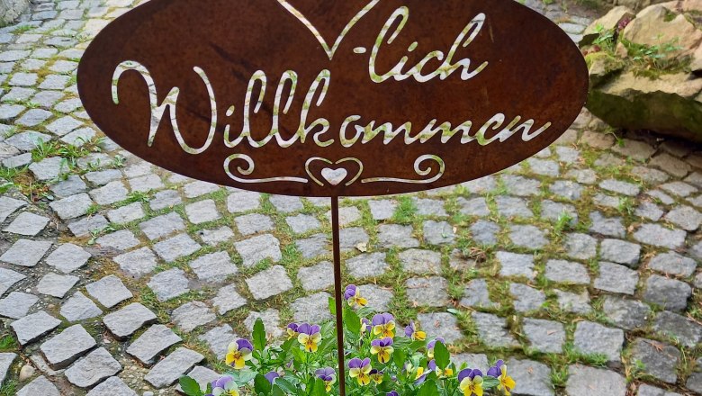 Herzlich Willkommen, © Familie Fleischmann Metallschild mit 'Herzlich Willkommen' über einem Topf mit gelb-violetten Blumen auf einem gepflasterten Weg.