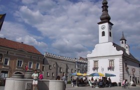Stadtplatz, © Gerold Guttmann Ein Stadtplatz mit einem weißen Gebäude mit Turm und Uhr, Menschen sitzen unter Sonnenschirmen, ein Brunnen im Vordergrund.