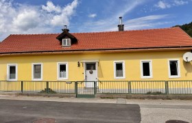 Hausansicht, © Christoph Menhofer Gelbes Haus mit rotem Dach und mehreren Fenstern, umgeben von einem grünen Zaun.