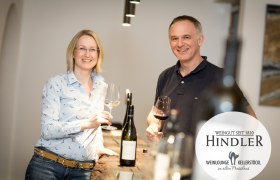 Rosi und Karl Hindler, © Weingut Hindler Ein Winzerpaar steht lächelnd mit Weingläsern in der Hand an einem Tisch im Weingut Hindler.