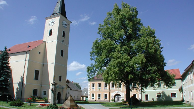 Hirschbach, © Hirschbach Kirche mit Turm und Uhr in einem Dorfplatz mit Bäumen und Gebäuden.