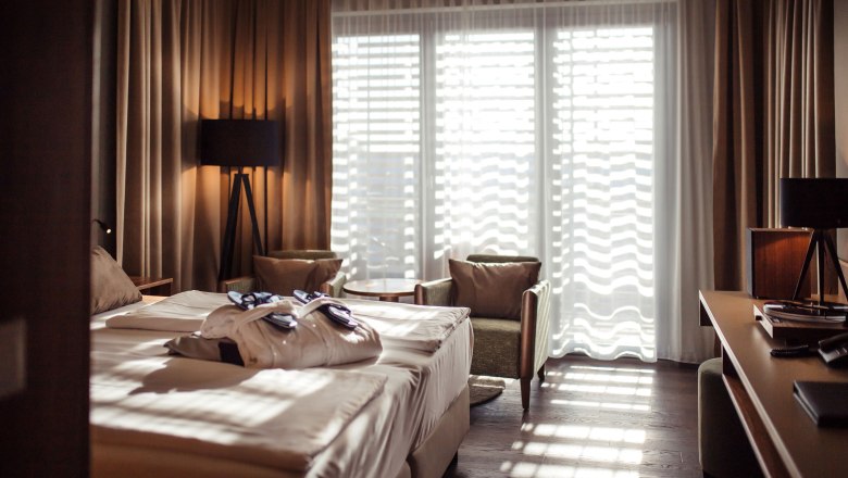 SILENT CLUB Zimmer der ThermeLaa, © Therme Laa Hotelzimmer mit Doppelbett, Bademänteln, zwei Sesseln und einem Tisch vor einem großen Fenster mit Vorhängen.