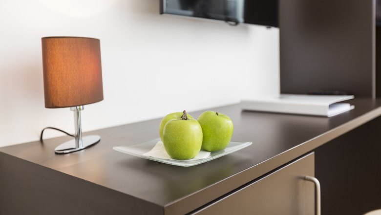 Gästezimmer Jöchlinger, © Ernst Jöchlinger Ein Tisch mit drei grünen Äpfeln auf einem Teller, einer Lampe und einem Fernseher im Hintergrund.