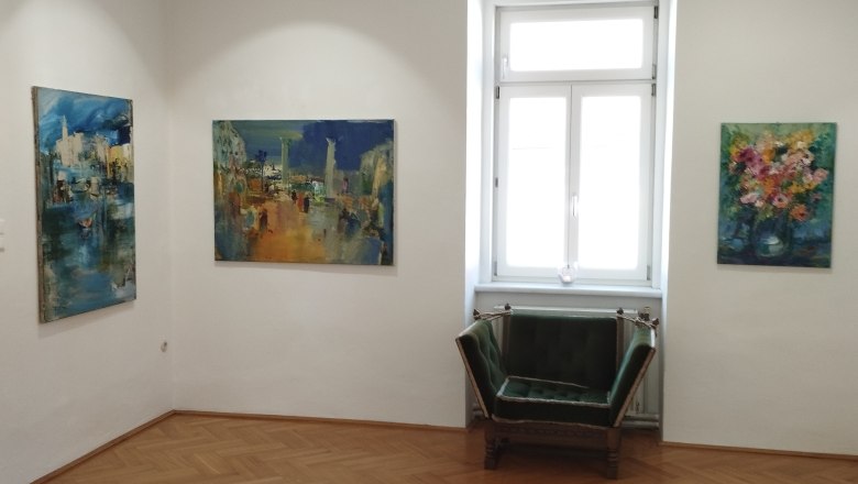 die Galerie MERKIMA, © Doris Hauser Innenansicht einer Galerie mit drei Gemälden an den Wänden und einem grünen Sessel vor einem Fenster.