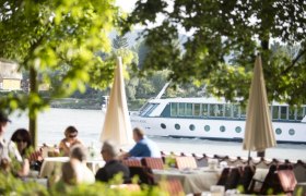 Blick auf die Donau, © Astrid Bartl Ein Flusskreuzfahrtschiff fährt auf der Donau, im Vordergrund sitzen Menschen in einem Café unter Bäumen.