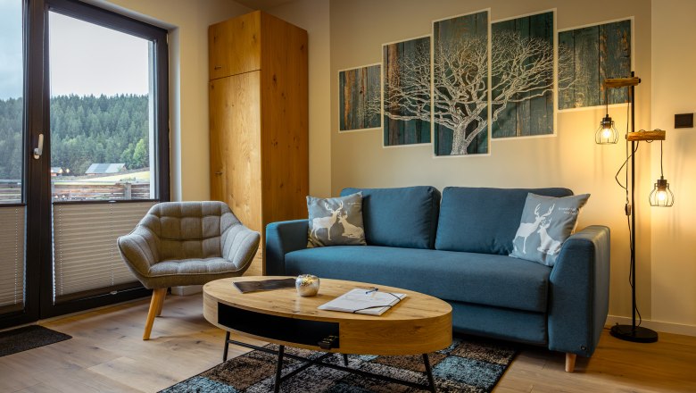 Ötscher Lodge, © Niederösterreich Werbung / Maximilian Pawlikowsky Gemütliches Wohnzimmer mit blauer Couch, Sessel, Holztisch und Waldblick durch Fenster.