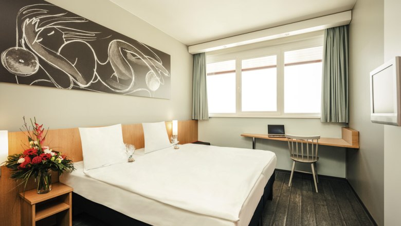 Doppelzimmer, © Marché Restaurants Österreich GmbH Modernes Doppelzimmer mit großem Bett, Kunstwerk an der Wand, Schreibtisch und Blumenarrangement.