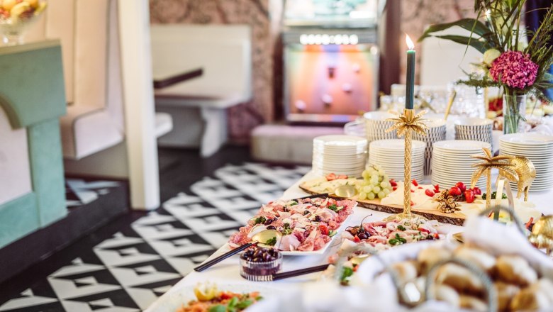 Buffet, © Matthias Kronfuss Ein reichhaltiges Buffet mit verschiedenen Speisen, Geschirrstapeln und dekorativen Kerzenhaltern in einem stilvollen Raum.