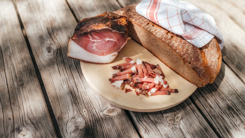 Selbstgemachtes aus der eigenen Landwirtschaft, © Niederösterreich Werbung/Daniela Führer Ein Holzbrett mit Brot, Speck und Speckstreifen auf einem Holztisch.