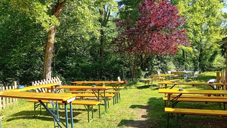 TDR-Zur MUCKE Garten, © B.Grohotolszky Ein Biergarten mit leeren Holzbänken und Tischen unter Bäumen.